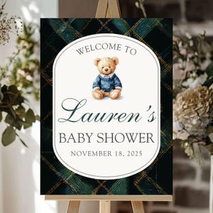 Pode incluir: Uma placa de chá de bebê em um cavalete. A placa tem um fundo xadrez com um oval branco no centro. O oval tem um ursinho de pelúcia com um suéter azul e o texto "Welcome to Lauren's Baby Shower November 18, 2025."