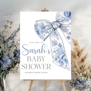 May include: A baby shower sign featuring a large blue and white floral bow. The sign reads "Welcome to Sarah's Baby Shower" with the date "Saturday March 24 20XX". The sign is displayed on a wooden easel.