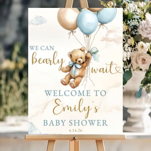 Puede incluir: Un letrero de baby shower sobre un caballete presenta una ilustración de acuarela de un osito de peluche sosteniendo globos. El letrero dice "We can bearly wait" y "Welcome to Emily's Baby Shower" con la fecha 4.14.26. La combinación de colores incluye beige, azul y dorado.