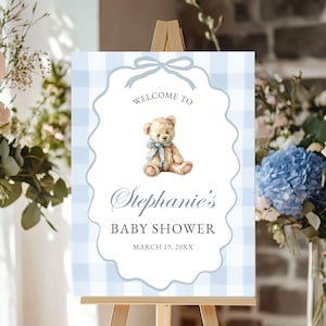 May include: A baby shower sign on a wooden easel. The sign has a blue and white gingham pattern background with a decorative border. The sign reads "Welcome to Stephanie's Baby Shower" with a teddy bear illustration and the date.