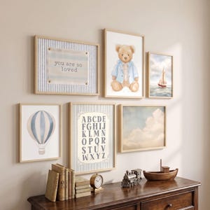 May include: A gallery wall arrangement featuring six framed prints: a striped sign with the text you are so loved, a teddy bear in a blue jacket, a sailboat on water, a hot air balloon, an alphabet chart, and a cloud-filled sky, above a wooden dresser.