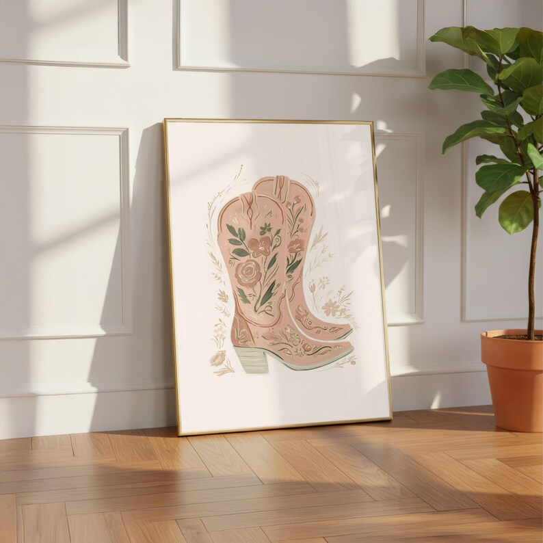 May include: A framed wall art print featuring a pair of light pink cowboy boots decorated with intricate green and brown floral illustrations. The boots are surrounded by small, delicate botanical sketches on a plain, off-white background.