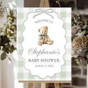 May include: A baby shower welcome sign on a wooden easel. The sign features a watercolor teddy bear illustration and text that reads "Welcome to Stephanie's Baby Shower" with the date March 19, 20XX. The background has a light green and white gingham pattern.