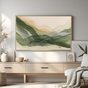 Op de afbeelding: Abstracte aquarel landschapskunst in een houten lijst. Het kunstwerk toont gelaagde groene en bruine tinten, met witte lineaire accenten. De ingelijste kunst wordt tentoongesteld boven een licht houten kast, met andere decoratieve items.