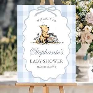 May include: A baby shower welcome sign featuring a light blue gingham pattern background. The sign includes the text "Welcome to" above an illustration of Winnie the Pooh with a honey pot. The sign reads "Stephanie's BABY SHOWER" and the date.