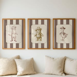 May include: Three framed wall art prints featuring brown ink illustrations of a cat, a teddy bear, and a goose, each wearing a cowboy hat. The prints are set against a background of vertical brown and cream stripes within a light wood frame.