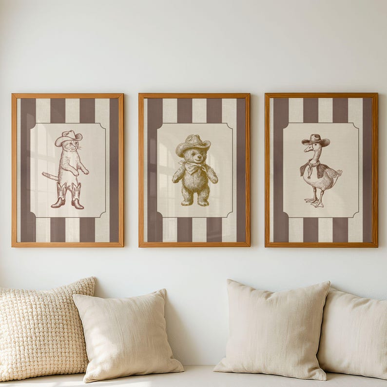 May include: Three framed wall art prints featuring brown ink illustrations of a cat, a teddy bear, and a goose, each wearing a cowboy hat. The prints are set against a background of vertical brown and cream stripes within a light wood frame.