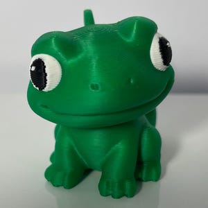 May include: A vibrant green frog ornament with large, cartoonish eyes. The frog has a textured finish and is depicted in a seated posture. The eyes are white with black pupils. A small, rounded projection is on the top of its head.