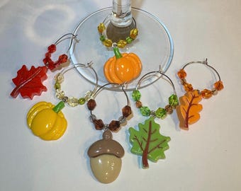 Puede incluir: Un conjunto de adornos para copas de vino con temática otoñal. Los adornos presentan varias formas, incluyendo calabazas, hojas y una bellota. Los adornos son en tonos naranja, amarillo, rojo y verde, con detalles de cuentas y aros plateados.