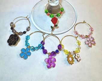 May include: Set of six colorful glass charms with flower and rose designs. Each charm has a gold ring and is decorated with beads. The charms are perfect for identifying wine glasses at a party.