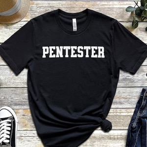 Puede incluir: Camiseta negra con la palabra "PENTESTER" impresa en letras blancas en negrita. La camiseta se muestra sobre una superficie de madera, junto con un par de zapatillas deportivas blancas y negras, un sombrero de paja y gafas.