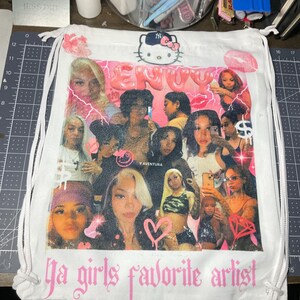 May include: White drawstring bag with a pink and white graphic print of a collage of women's faces, a pink heart, and the text "Ja girls favorite artist". The bag also features a pink Hello Kitty graphic.