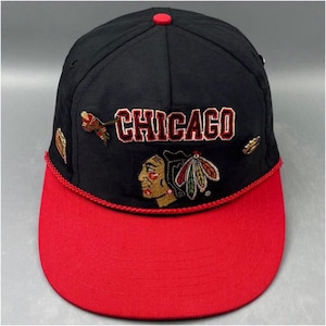 Peut inclure: Une casquette de baseball noire avec une visière rouge et une bordure en corde rouge. L'avant présente le mot CHICAGO en lettres brodées rouges au-dessus d'un logo brodé représentant le profil d'une personne avec des plumes.