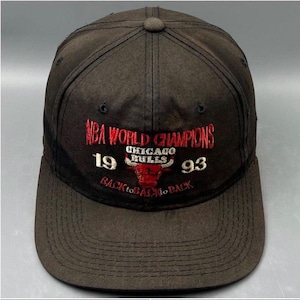 Op de afbeelding: Een zwarte honkbalpet met geborduurd rood en wit tekstontwerp met de tekst NBA WORLD CHAMPIONS 1993 BACK TO BACK, inclusief een centraal Chicago Bulls logo met een rode stierenkop boven de teamnaam in witte letters.
