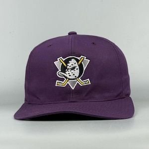 Può includere: Un cappellino da baseball viola con un logo ricamato sul davanti. Il logo raffigura una maschera da hockey bianca e nera a forma di testa di papero, centrata sopra due mazze da hockey gialle incrociate su uno sfondo triangolare grigio.