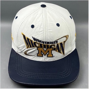 May include: A white baseball cap with a dark blue brim. The front features embroidered text that reads MICHIGAN WOLVERINES with a large gold letter M. The cap has dark blue eyelets and a dark blue button at the top centre of the crown.