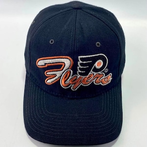 Peut inclure: Une casquette de baseball noire avec le logo des Philadelphia Flyers brodé sur le devant. Le logo se compose du mot Flyers en lettres cursives orange et blanches, surmonté d'un emblème stylisé en forme de P ailé noir et orange.