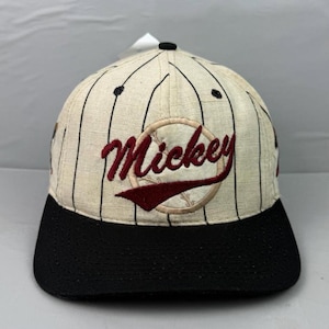 Op de afbeelding: Een honkbalpet met een crèmekleurige kroon met dunne zwarte krijtstrepen en een effen zwarte klep. De voorkant is voorzien van een geborduurde ronde patch met een honkbalafbeelding en het woord Mickey in donkerrood cursief schrift.