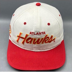 Könnte beinhalten: Eine weiße Baseballkappe mit rotem Schirm und einem roten Knopf oben. Auf der Vorderseite ist der Text Atlanta Hawks in Rot mit gelben Umrandungen gestickt. Ein kleines rotes Logo ist an der Seite zu sehen und rote Ösen sind um die Krone verteilt.