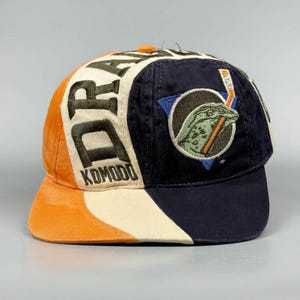 Op de afbeelding: Een veelkleurige baseballpet met een ontwerp van oranje, crèmekleurige en marineblauwe panelen. Het crèmekleurige paneel toont de geborduurde tekst "DRAGONS KOMODO" in donkergroen, terwijl het marineblauwe paneel een geborduurde hagedisafbeelding bevat.