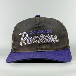 May include: A black baseball cap with a purple brim and a purple button on top. The front features the embroidered text Colorado Rockies in a white and purple script font. The cap has a structured crown with six panels and ventilation eyelets.
