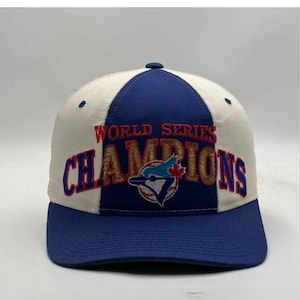 Op de afbeelding: Een honkbalpet in een blauw en wit kleurenschema. Het voorpaneel toont de geborduurde tekst WORLD SERIES CHAMPIONS in rode en gouden letters, met een logo van een blauwe gaai gecentreerd boven het woord CHAMPIONS.