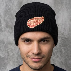 May include: A black ribbed knit beanie featuring an embroidered patch on the front cuff. The patch displays a red and white winged wheel logo. The hat is constructed from a thick, textured yarn material with a folded brim design.