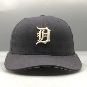 Op de afbeelding: Een donker marineblauwe baseballpet met een opvallende, geborduurde witte letter D in een gotisch lettertype, gecentreerd op het voorpaneel. De pet heeft een gestructureerde kroon, een gebogen klep en gestikte oogjes voor ventilatie.