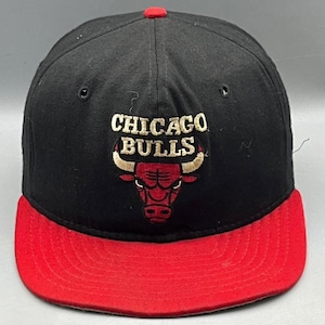 Op de afbeelding: Een zwarte honkbalpet met een rode platte klep en een rode knoop aan de bovenkant. De voorkant is voorzien van het geborduurde Chicago Bulls-logo in rood, wit en beige, met de tekst Chicago Bulls in beige boven de stierenkopafbeelding.