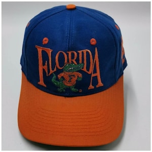 Può includere: Un cappellino da baseball blu con visiera arancione e un bottone arancione sulla parte superiore. La parte anteriore presenta la parola FLORIDA ricamata in lettere arancioni, con un alligatore verde che indossa una maglia arancione con una F.