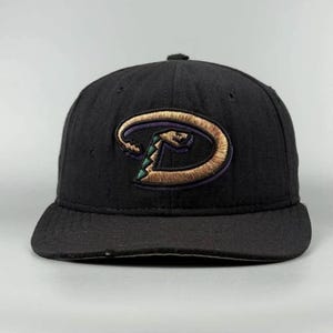 May include: A black baseball cap featuring an embroidered logo on the front. The logo is a stylised letter D in metallic gold thread with a dark purple outline and a small green snake-like accent integrated into the shape of the letter.