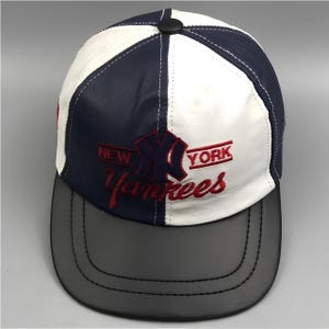 Op de afbeelding: Een baseballpet met een gespleten ontwerp in marineblauw en wit. De voorkant toont het geborduurde New York Yankees logo en de tekst in rood en marineblauw. De pet heeft een zwarte platte klep en een zwarte knoop op de bovenkant van de kroon.