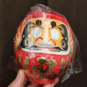 May include: A red Daruma doll with gold accents and Japanese calligraphy. The doll has a white face with black eyes and eyebrows, and a yellow and orange design around the eyes. The doll is wrapped in clear plastic.