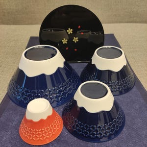 Pu&ograve; includere: Set di quattro ciotole in ceramica blu e bianca con un design a forma di montagna. Le ciotole sono annidate l'una dentro l'altra e sono decorate con un cappello di neve bianco e un design di montagna blu. La ciotola pi&ugrave; piccola &egrave; rossa.