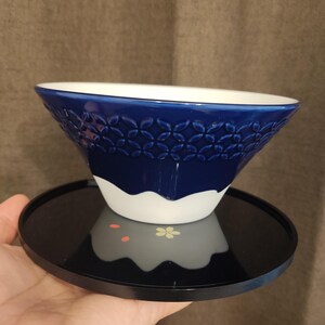 Pu&ograve; includere: Una ciotola in ceramica blu e bianca con un motivo decorativo. La ciotola &egrave; appoggiata su un vassoio nero.