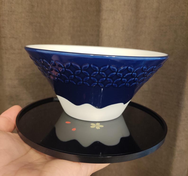 Pu&ograve; includere: Una ciotola in ceramica blu e bianca con un motivo decorativo. La ciotola &egrave; appoggiata su un vassoio nero.