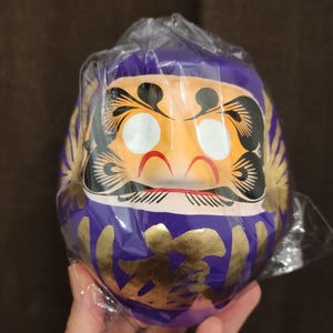 May include: A Daruma doll, a traditional Japanese good luck charm, is encased in plastic. The doll features a purple body, gold accents, and an orange face with black details. The doll has white eyes and red accents.