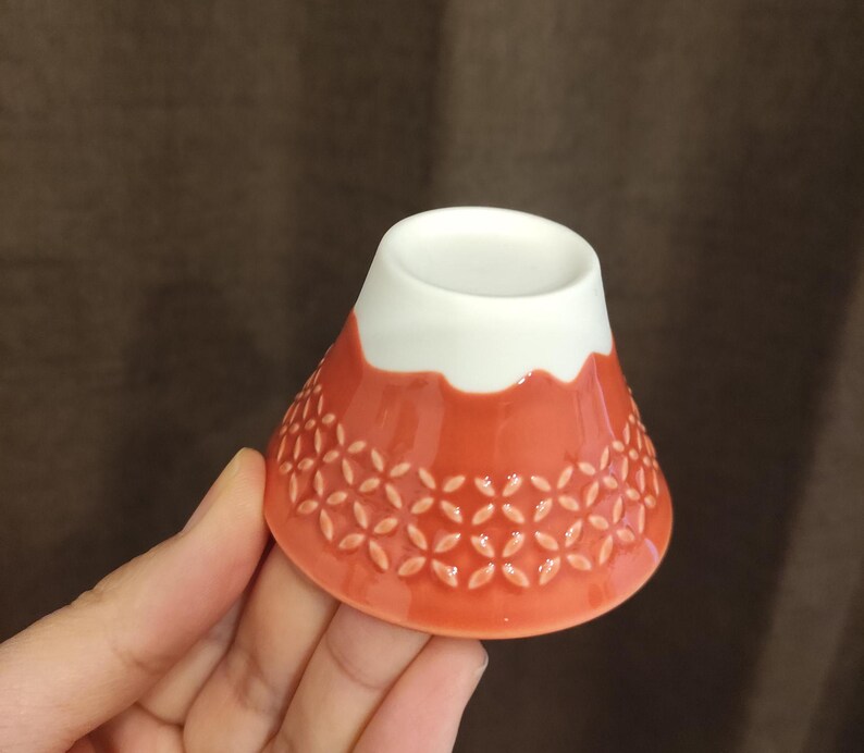 Pu&ograve; includere: Una piccola ciotola di ceramica rossa con un bordo bianco e un motivo floreale. La ciotola ha la forma di una montagna.