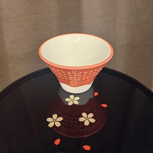 Pu&ograve; includere: Una piccola tazza da t&egrave; in ceramica arancione con interno bianco e un motivo floreale. La tazza &egrave; appoggiata su una superficie nera con un motivo floreale.