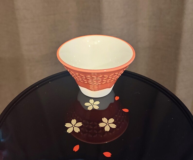 Pu&ograve; includere: Una piccola tazza da t&egrave; in ceramica arancione con interno bianco e un motivo floreale. La tazza &egrave; appoggiata su una superficie nera con un motivo floreale.