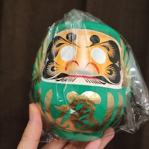 Könnte beinhalten: Eine grüne Daruma-Puppe mit goldenen Akzenten und einem stilisierten Gesicht, verpackt in klarem Kunststoff. Die Puppe hat einen roten Mund, weiße Augen und schwarze Details. Das japanische Schriftzeichen für "Glück" ist in Gold auf der Vorderseite geschrieben.