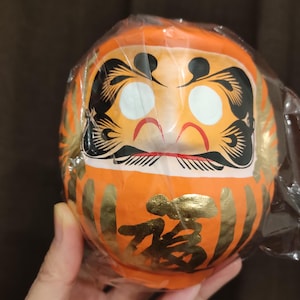 Puede incluir: Muñeca Daruma naranja con detalles faciales negros y detalles dorados, envuelta en plástico transparente. El carácter japonés para "fortuna" está escrito en oro en la parte delantera de la muñeca.