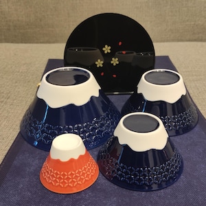 Pu&ograve; includere: Quattro ciotole in ceramica blu e bianca a forma di Monte Fuji con una ciotola rossa e bianca al centro. Le ciotole hanno un bordo bianco e un motivo blu e bianco.