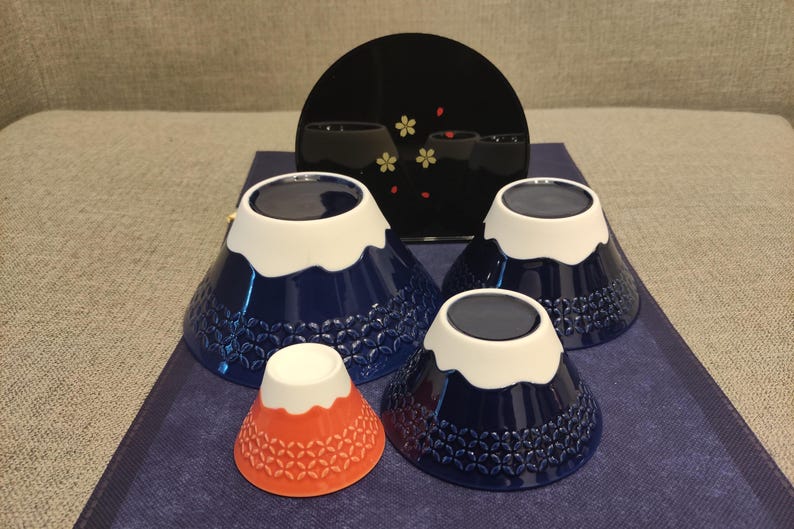 Pu&ograve; includere: Quattro ciotole in ceramica blu e bianca a forma di Monte Fuji con una ciotola rossa e bianca al centro. Le ciotole hanno un bordo bianco e un motivo blu e bianco.