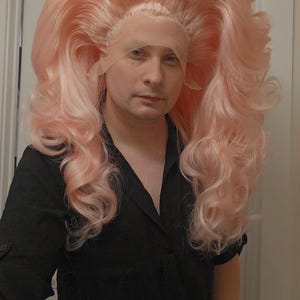 Może przedstawiać: Jasnoróżowa, obszerna peruka z długimi, kręconymi włosami. Peruka jest stylizowana z wysoką koroną i kaskadowymi lokami. Osoba na zdjęciu ma na sobie czarną koszulę. Peruka wydaje się być w stylu z koronkowym przodem.