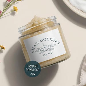 May include: A clear glass jar filled with a light-coloured scrub, featuring a white label with the text "EMA'S MOCKUPS" and a botanical illustration. A circular sticker with "INSTANT DOWNLOAD" is also visible. A sprig of flowers and a white towel are in the background.