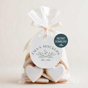 May include: A clear cellophane bag tied with a cream ribbon, filled with heart-shaped cookies. Each cookie has a white icing heart. A round tag with the text "EMA'S MOCKUPS" and "INSTANT DOWNLOAD" is attached to the bag.