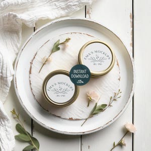 May include: Two gold-rimmed jars with white labels featuring the text "EMA'S MOCKUPS" and a botanical design, resting on a distressed white tray. The tray is adorned with sprigs of flowers and greenery, and a white cloth. A dark teal circle with the words "INSTANT DOWNLOAD" is also present.