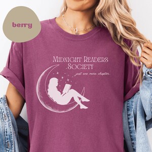 Könnte beinhalten: Ein maroonfarbenes T-Shirt mit einer weißen Grafik einer Frau, die auf einem Halbmond ein Buch liest. Der Text "Midnight Readers Society" und "Just one more chapter" ist auf dem Shirt aufgedruckt.