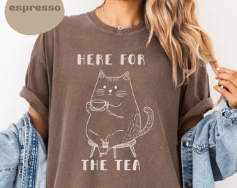 Peut inclure: Un t-shirt marron avec un motif blanc d'un chat assis sur une chaise et tenant une tasse. Le texte "Here for the tea" est imprimé sous le chat.